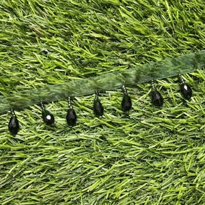 Può includere: Una striscia di nastro in velluto verde oliva con una fila di perline nere pendenti a forma di goccia. Ogni perlina è fissata al nastro da un piccolo anello metallico argentato e da una serie di minuscole perline scure, creando una frangia.