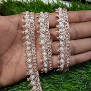 Peut inclure: Gros plan d'une garniture en dentelle rose gold avec des perles blanches. La garniture est délicate et complexe, avec un motif répétitif de petites fleurs et de feuilles. Les perles sont uniformément espacées le long du bord de la garniture.