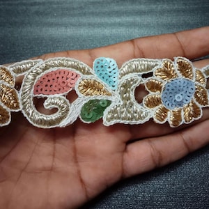 May include: Close-up of a decorative trim with floral and scroll designs. The trim features gold, white, pink, blue, and green sequins and embroidery. The trim is held in a hand.