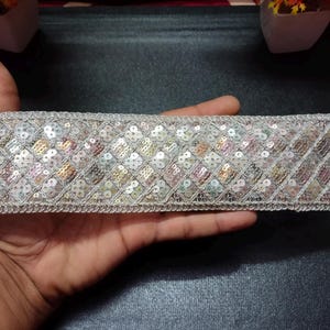 May include: A silver sequined trim with a geometric pattern. The trim is made of small, iridescent sequins that are sewn onto a white fabric base.