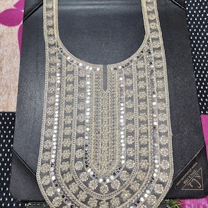 Może przedstawiać: Beżowy, haftowany dekolt z kwiatowymi i okrągłymi lustrzanymi zdobieniami. Projekt ma kształt litery U, z centralnym pionowym panelem i ozdobnymi obramowaniami. Element jest prezentowany na czarnej powierzchni.