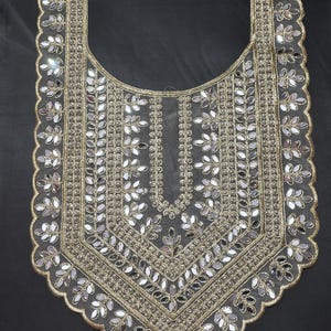 May include: A decorative, gold-toned embroidered neckline with a scalloped edge. The design features mirrored accents and leaf-like patterns, creating a shimmering effect. The neckline is set against a dark background, highlighting the intricate details.