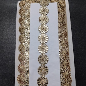 Può includere: Passamaneria in pizzo floreale dorato su supporto bianco. La passamaneria presenta un motivo ripetuto di fiori traforati, adatta per abbellire capi di abbigliamento o lavori artigianali. La passamaneria è larga circa 2,5 cm.