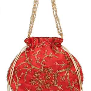 May include: A round red drawstring pouch featuring gold floral embroidery. The bag includes a handle made of white beads and gold-toned accents, with two side drawstrings finished with small gold-toned bell-shaped tassels at the ends.