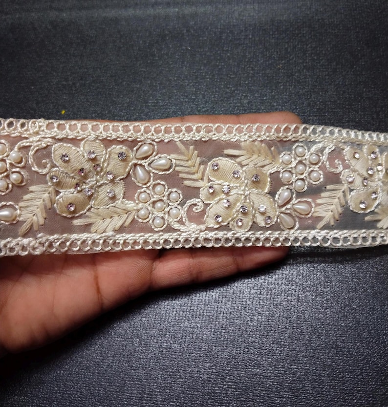 May include: White beaded lace trim with pearl accents and floral embroidery. The trim is delicate and intricate, perfect for adding a touch of elegance to any project.