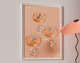Könnte beinhalten: Ein gerahmter Druck von vier Champagnerflöten mit einem pfirsichfarbenen Hintergrund. Die Flöten sind mit einer klaren Flüssigkeit gefüllt und haben jeweils ein kleines blaues Objekt, das darin schwimmt. Im Vordergrund steht ein rosa Lampenschirm mit einem schwarzen Standfuß.