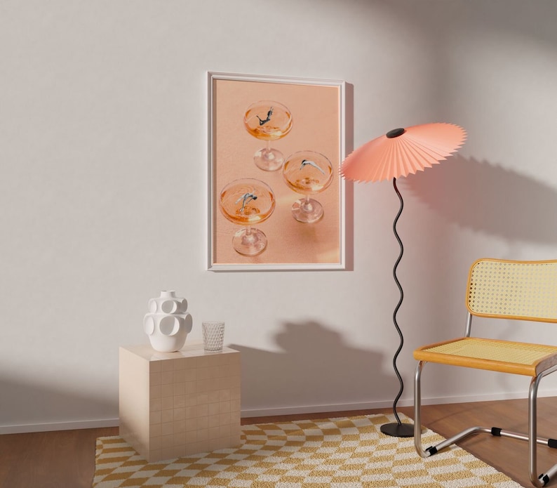 May include: A framed print of four champagne flutes with a peach background. The flutes are filled with a clear liquid and have a small blue object floating in each. A pink lampshade with a black stand is in the foreground.