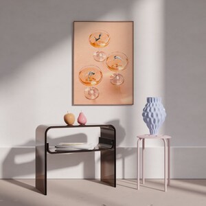 May include: A framed print of a still life featuring four champagne glasses with figures diving into the liquid. The print is hanging on a white wall above a brown side table with two small decorative objects. A pink stool with a blue ceramic vase sits in the foreground.