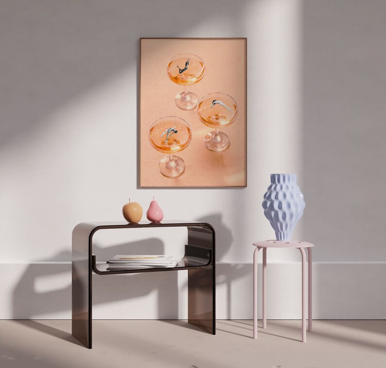 May include: A framed print of a still life featuring four champagne glasses with figures diving into the liquid. The print is hanging on a white wall above a brown side table with two small decorative objects. A pink stool with a blue ceramic vase sits in the foreground.