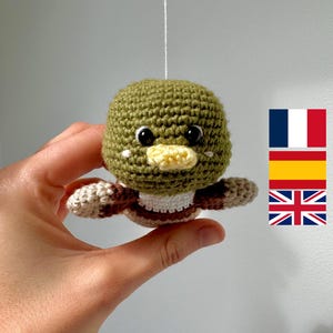 May include: A green crocheted stuffed animal with a yellow beak and brown and white stripes. The animal is hanging from a white string.