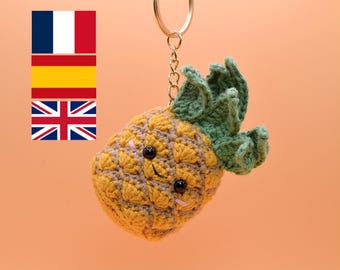 May include: A handmade crochet pineapple keychain with a gold-colored keyring. The pineapple is yellow and gray with green leaves and black eyes. The keychain is suspended against a peach-colored background.