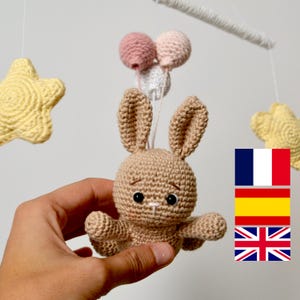 May include: A handmade crochet baby mobile featuring a tan bunny with black eyes and a small pink nose, holding three pink and white balloons. Two yellow stars hang on either side. The mobile is designed for a nursery.