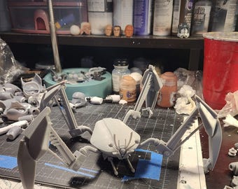 May include: A light gray, unpainted plastic model of a multi-legged mechanical creature with a central body and four articulated, angular limbs. The figure features a segmented head with thin antennae and is positioned on a black cutting mat.