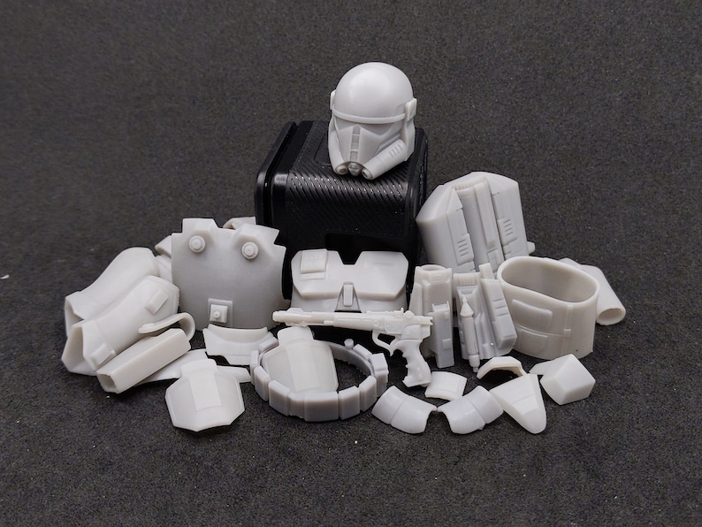 K&ouml;nnte beinhalten: Eine Sammlung grauer Kunststoff-Modellbausatzteile, darunter ein Helm, eine Brustpanzerung und verschiedene andere Komponenten. Die Teile sind auf einer dunklen Oberfl&auml;che angeordnet, was auf ein Hobby- oder Bastelprojekt hindeutet. Das Set scheint f&uuml;r eine Science-Fiction-Figur zu sein.