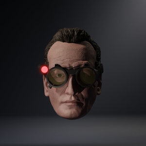 May include: A close-up of a man's face wearing round, dark-rimmed goggles with a red light on the side. The goggles have a yellow tint to the lenses.