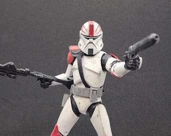 May include: A white and red action figure of a clone trooper, holding a blaster rifle and a pistol. The figure has a weathered appearance, with red accents on the helmet and armor. The figure is posed in a dynamic stance against a dark background.