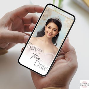 May include: A smartphone displays a wedding invitation with the text "Save the Date." The invitation features a smiling woman wearing a tiara and a light pink dress. The background is a soft blue with floral accents. The phone is held in two hands.