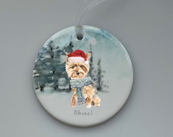May include: A round ceramic ornament featuring a watercolor illustration of a Yorkshire Terrier wearing a Santa hat and a blue scarf. The ornament has a white background with a snowy forest scene and the text "[Name]" at the bottom.