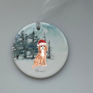May include: A round ceramic bauble featuring a watercolour illustration of a dog wearing a Santa hat in a snowy woodland setting. The bauble has a white ribbon for hanging and the text "[Name]" at the bottom.