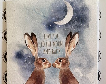 Puede incluir: Una tarjeta de felicitación con una ilustración de acuarela de dos conejos marrones mirándose bajo una luna creciente. La tarjeta tiene el texto "Love you to the moon and back" en letras doradas.