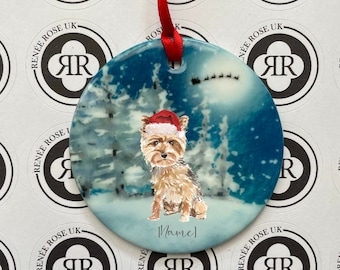 May include: A round, blue and white Christmas ornament with a painted image of a Yorkshire Terrier wearing a Santa hat. The dog is sitting in a snowy forest scene. The ornament has the text "[Name]" written on it.