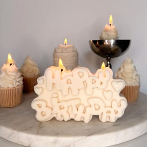 May include: A cream-colored candle shaped like the words HAPPY BIRTHDAY with two lit wicks, surrounded by several smaller cupcake-shaped candles with lit wicks, including one in a silver pedestal bowl, all arranged on a round white marble surface.