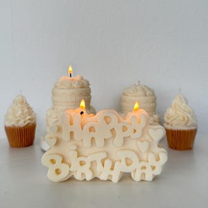 May include: An off-white candle shaped like the words Happy Birthday with small heart accents, featuring two lit wicks. Behind it are two off-white cake-shaped candles and two off-white cupcake-shaped candles in brown paper liners, all lit.