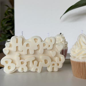 May include: An off-white wax candle shaped like a rectangular plaque with the embossed text "HAPPY BIRTHDAY" and small heart accents, accompanied by two smaller cupcake-shaped candles with textured frosting tops and tan ribbed bases.