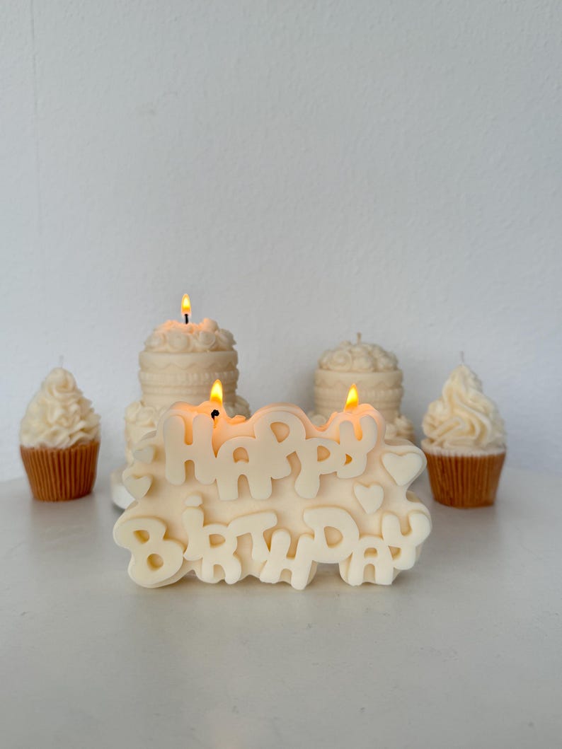 May include: An off-white candle shaped like the words Happy Birthday with small heart accents, featuring two lit wicks. Behind it are two off-white cake-shaped candles and two off-white cupcake-shaped candles in brown paper liners, all lit.