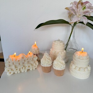 May include: A collection of off-white wax candles shaped like a Happy Birthday sign, two tiered cakes, and two cupcakes. The Happy Birthday sign and one cupcake are lit with small flames. The candles feature detailed frosting-like textures.