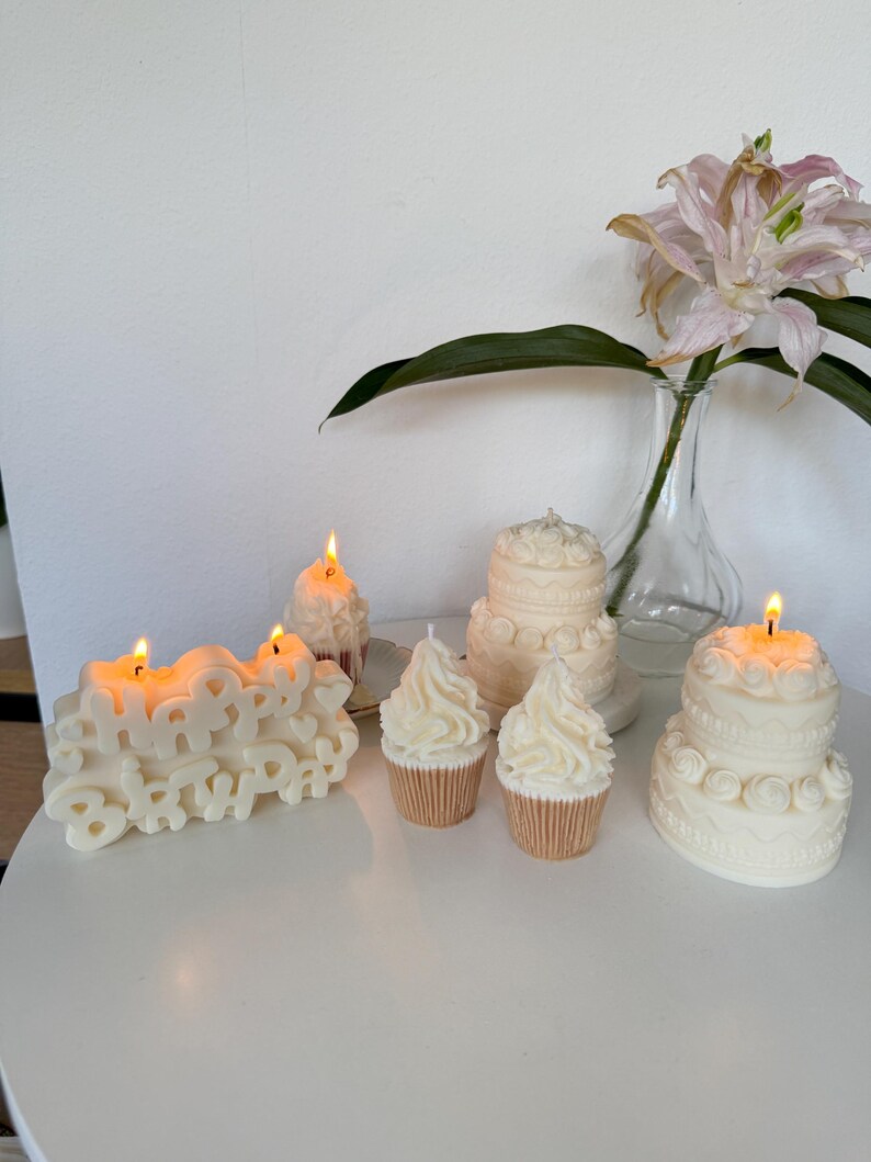 May include: A collection of off-white wax candles shaped like a Happy Birthday sign, two tiered cakes, and two cupcakes. The Happy Birthday sign and one cupcake are lit with small flames. The candles feature detailed frosting-like textures.
