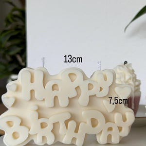 May include: An off-white wax candle shaped like the words "HAPPY BIRTHDAY" in a bubbly font, featuring small heart accents. The candle measures 13 cm in width and 7.5 cm in height, with two visible white wicks protruding from the top surface.