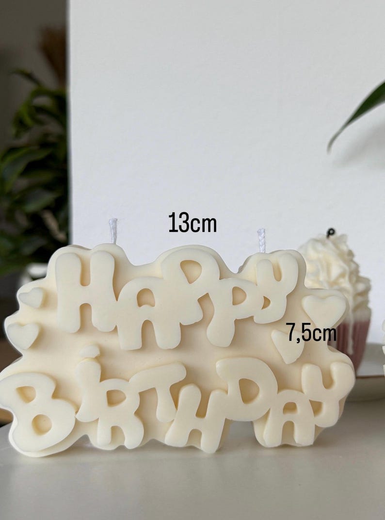 May include: An off-white wax candle shaped like the words "HAPPY BIRTHDAY" in a bubbly font, featuring small heart accents. The candle measures 13 cm in width and 7.5 cm in height, with two visible white wicks protruding from the top surface.