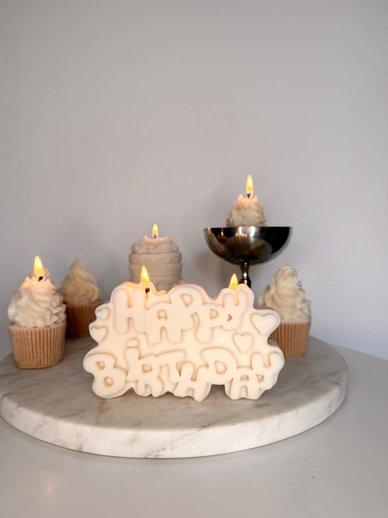 May include: A cream-colored candle shaped like the words HAPPY BIRTHDAY with two lit wicks, surrounded by several smaller cupcake-shaped candles with lit wicks, including one in a silver pedestal bowl, all arranged on a round white marble surface.
