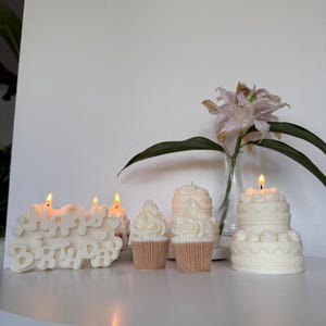 May include: A collection of white wax candles shaped like a Happy Birthday sign, two cupcakes with swirled frosting, and a two-tiered cake with decorative piping. Each candle is lit, casting a warm glow, and arranged on a white surface.