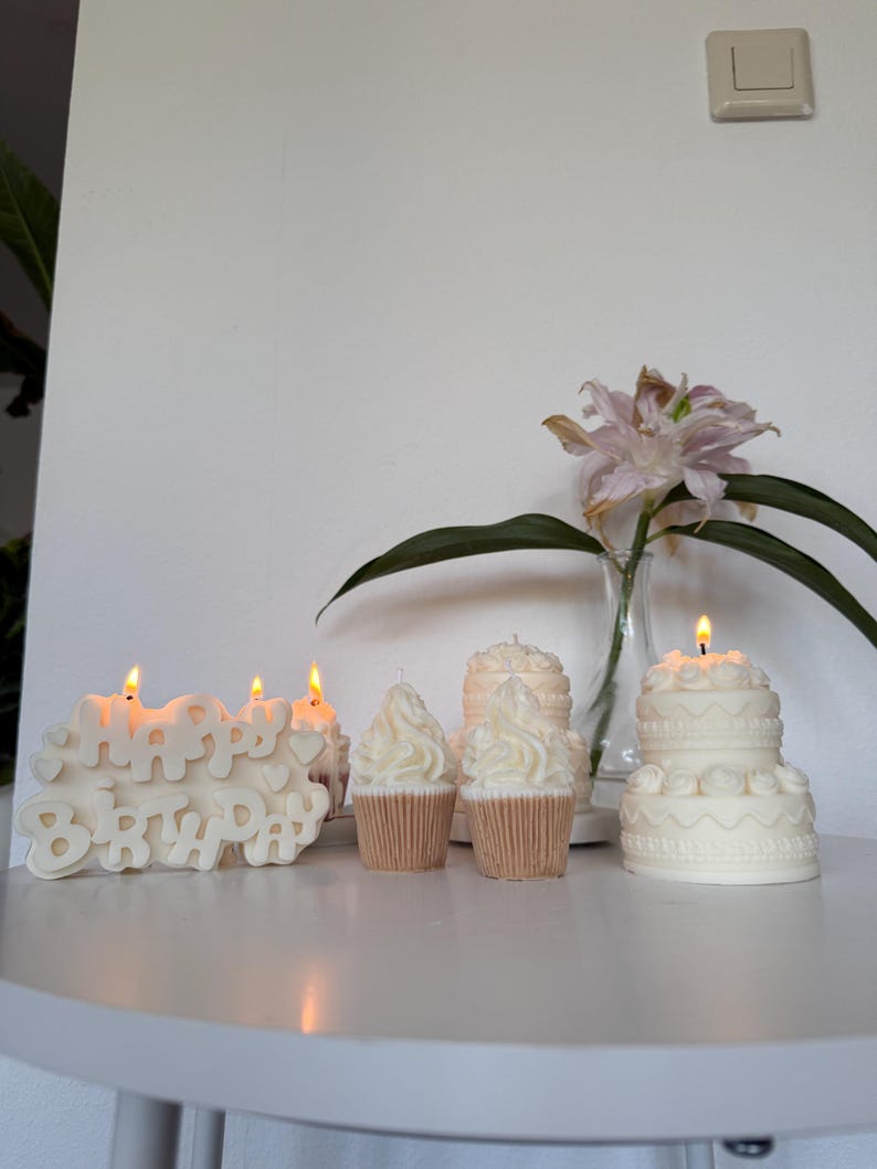 May include: A collection of white wax candles shaped like a Happy Birthday sign, two cupcakes with swirled frosting, and a two-tiered cake with decorative piping. Each candle is lit, casting a warm glow, and arranged on a white surface.