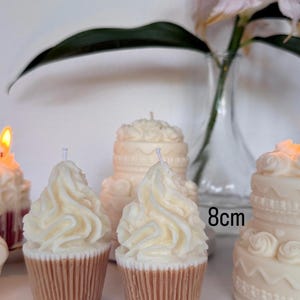May include: Two cupcake-shaped candles with white swirled wax frosting and tan ribbed bases, positioned in front of two larger tiered cake-shaped candles. One of the larger candles is labeled with the text 8cm, indicating its height.