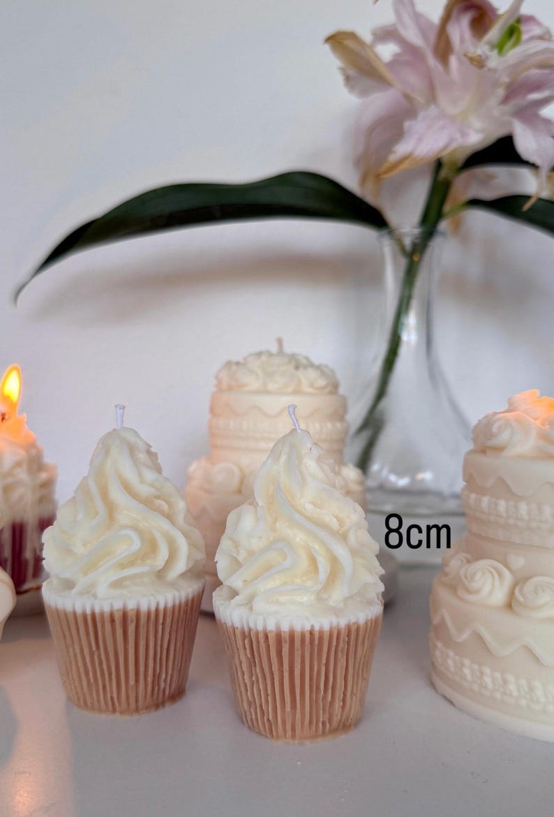 May include: Two cupcake-shaped candles with white swirled wax frosting and tan ribbed bases, positioned in front of two larger tiered cake-shaped candles. One of the larger candles is labeled with the text 8cm, indicating its height.