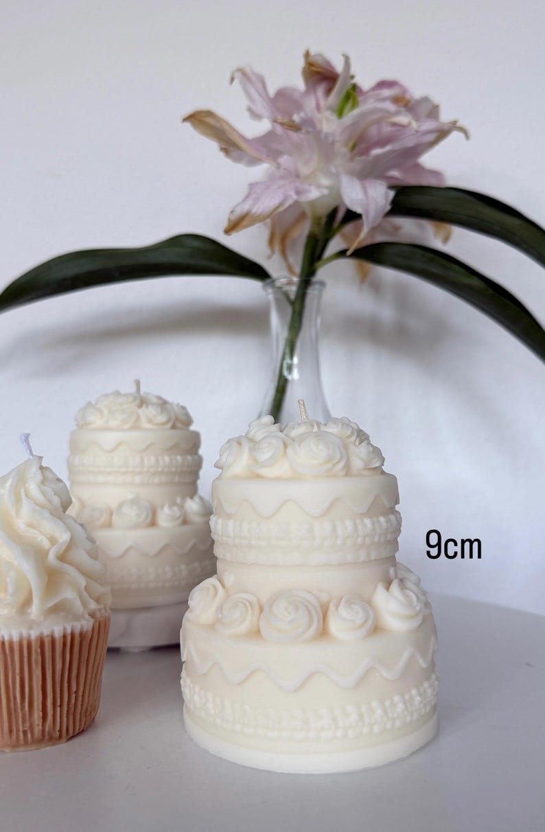 May include: Two off-white, two-tiered cake-shaped candles featuring intricate piped rose details, scalloped borders, and textured bead patterns. Each candle stands 9cm tall and includes a single centered wick at the top of the floral decoration.