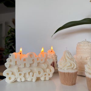 May include: A cream-colored candle shaped like the words HAPPY BIRTHDAY with small heart accents and two lit wicks. Beside it are two cupcake-shaped candles with swirled frosting tops and a small tiered cake candle in the background.