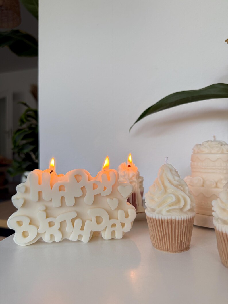 May include: A cream-colored candle shaped like the words HAPPY BIRTHDAY with small heart accents and two lit wicks. Beside it are two cupcake-shaped candles with swirled frosting tops and a small tiered cake candle in the background.