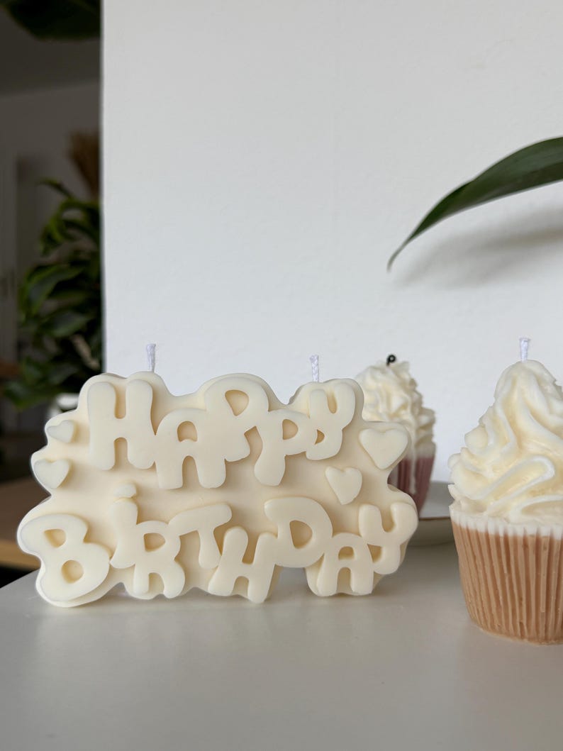 May include: An off-white wax candle shaped like a rectangular plaque with the embossed text "HAPPY BIRTHDAY" and small heart accents, accompanied by two smaller cupcake-shaped candles with textured frosting tops and tan ribbed bases.