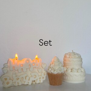 May include: A set of three off-white candles including a rectangular candle with the text Happy Birthday and two lit wicks, a cupcake-shaped candle with a tan base, and a two-tiered cake-shaped candle with decorative frosting details.
