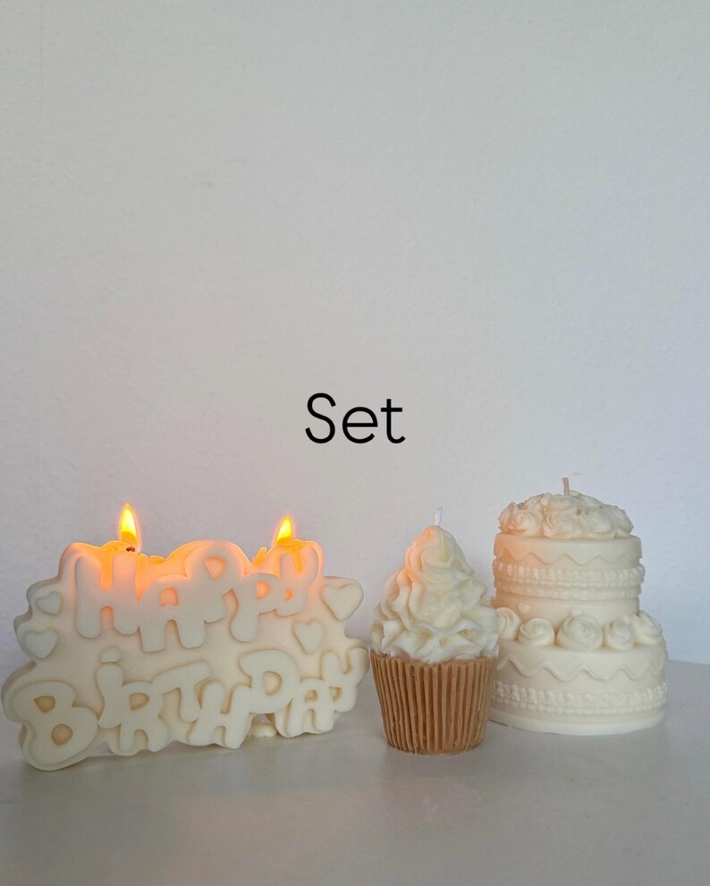 May include: A set of three off-white candles including a rectangular candle with the text Happy Birthday and two lit wicks, a cupcake-shaped candle with a tan base, and a two-tiered cake-shaped candle with decorative frosting details.