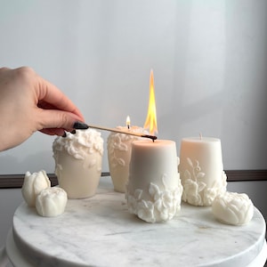 Könnte beinhalten: Eine Sammlung weißer Kerzen und blütenförmiger Wachsstücke. Einige Kerzen sind angezündet, mit sichtbaren Flammen. Die Kerzen sind mit floralen Reliefmustern verziert. Die Artikel sind auf einer weißen Marmoroberfläche angeordnet.