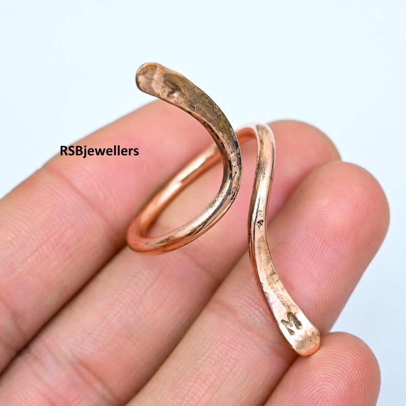 May include: A copper ring with a twisted, wavy design. The ring is worn on a finger and has a small 'M' stamped on the side.