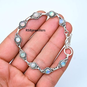 May include: A silver bracelet featuring oval opal gemstones set in a bezel design. The bracelet has a toggle clasp and is held in a hand. The text "RSBjewellers" is visible on the bracelet.
