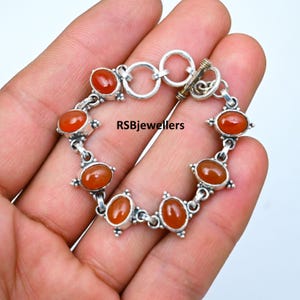 Puede incluir: Un brazalete de plata con cierre de palanca presenta siete piedras ovaladas de cornalina. El brazalete está hecho de plata de ley y tiene un acabado pulido. RSBjewellers está impreso en la imagen.