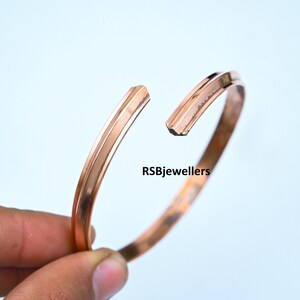 May include: A simple copper bangle bracelet with a slightly rounded shape. The bracelet is open and has a gap where the ends meet.