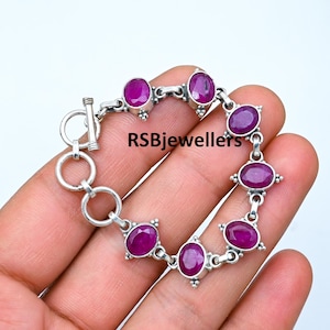 May include: A silver bracelet with a toggle clasp features a chain design with seven oval-shaped pink gemstones. The bracelet is on a hand, and the text "RSBjewellers" is visible in the background.